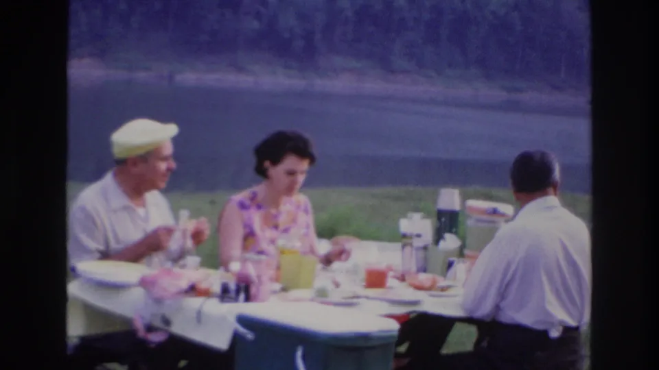 1967 Ottawa home-movie frame showing three adults eating at an outdoor picnic table, with a cooler in front and water behind them.