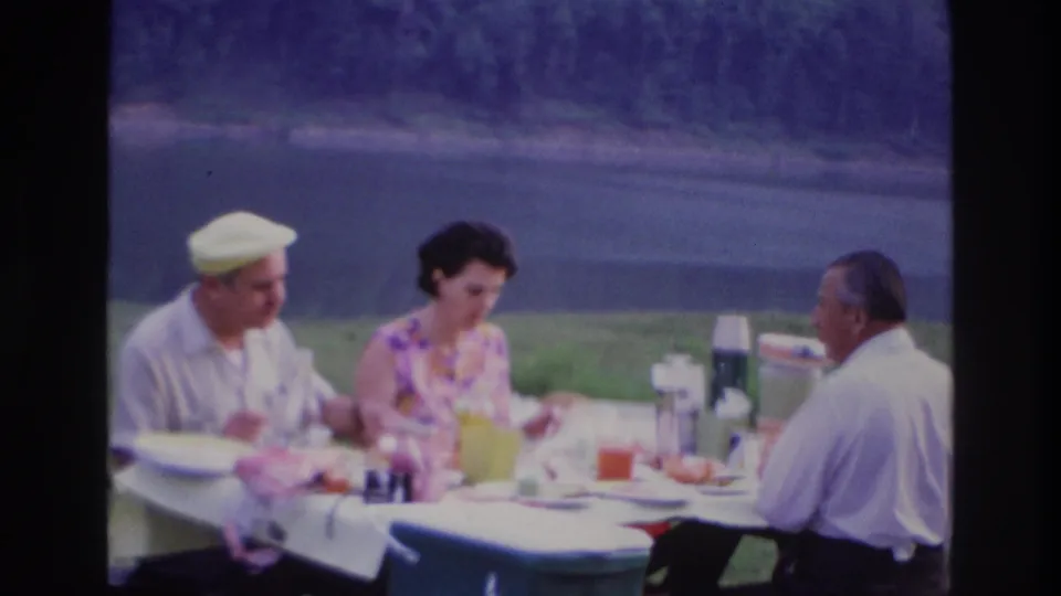 1967 Ottawa picnic frame with three adults seated at a table, food and drinks spread across a white cloth, and water visible beyond the grass.