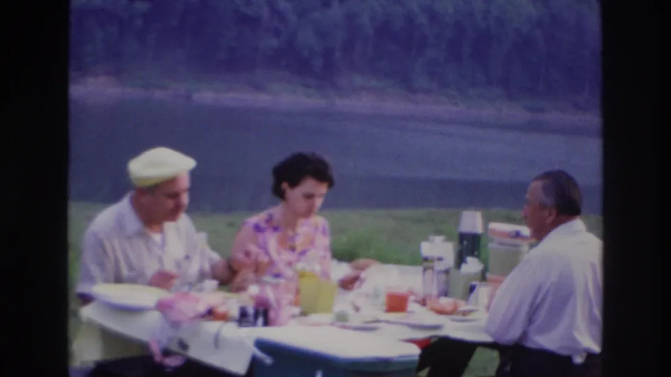 In Ottawa, Canada in 1967, three adults sit at a white-covered picnic table by a lake with plates, cups, and thermoses.