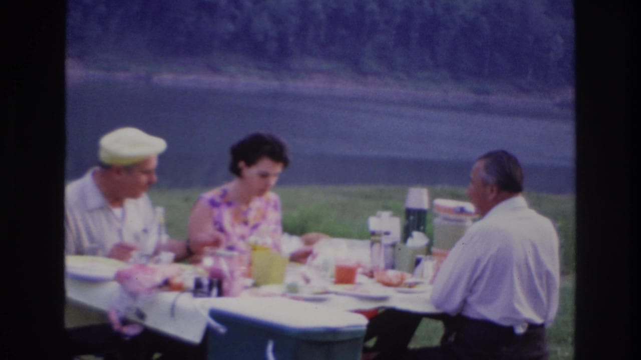 Three adults in 1960s attire eat and converse at an outdoor picnic table near water in Ottawa, Canada.