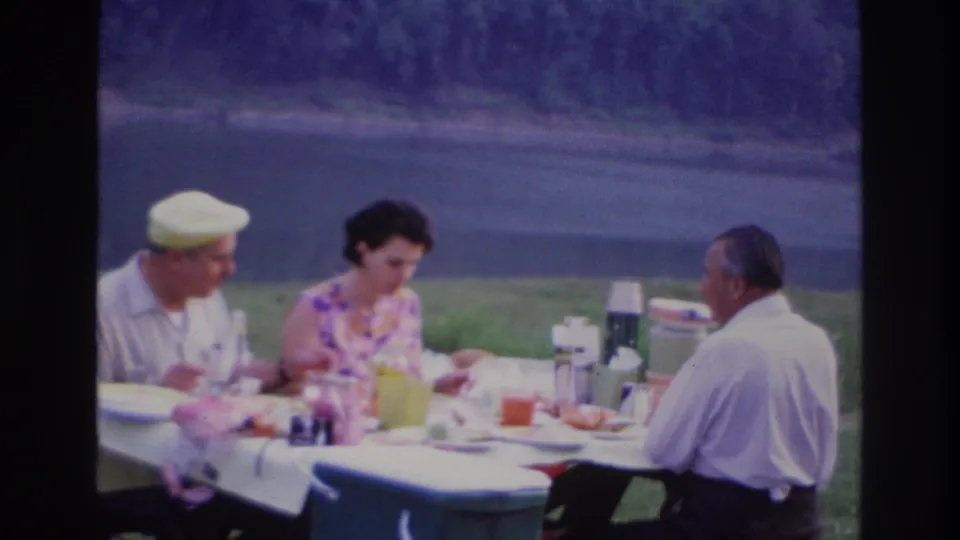 Three adults in 1960s attire eat and converse at an outdoor picnic table near water in Ottawa, Canada.