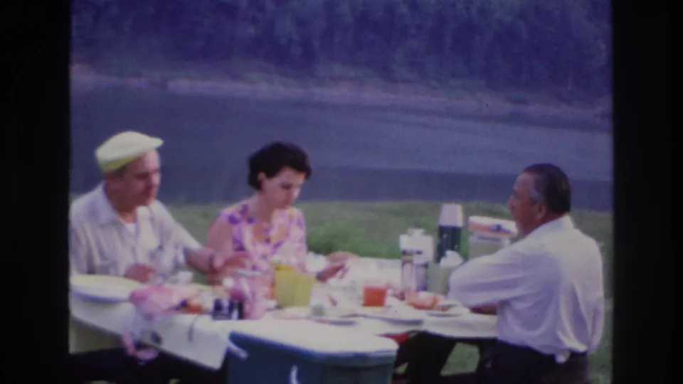 A 1967 home-movie frame shows three adults at a picnic table with food, cups, and thermoses beside a lake in Ottawa.