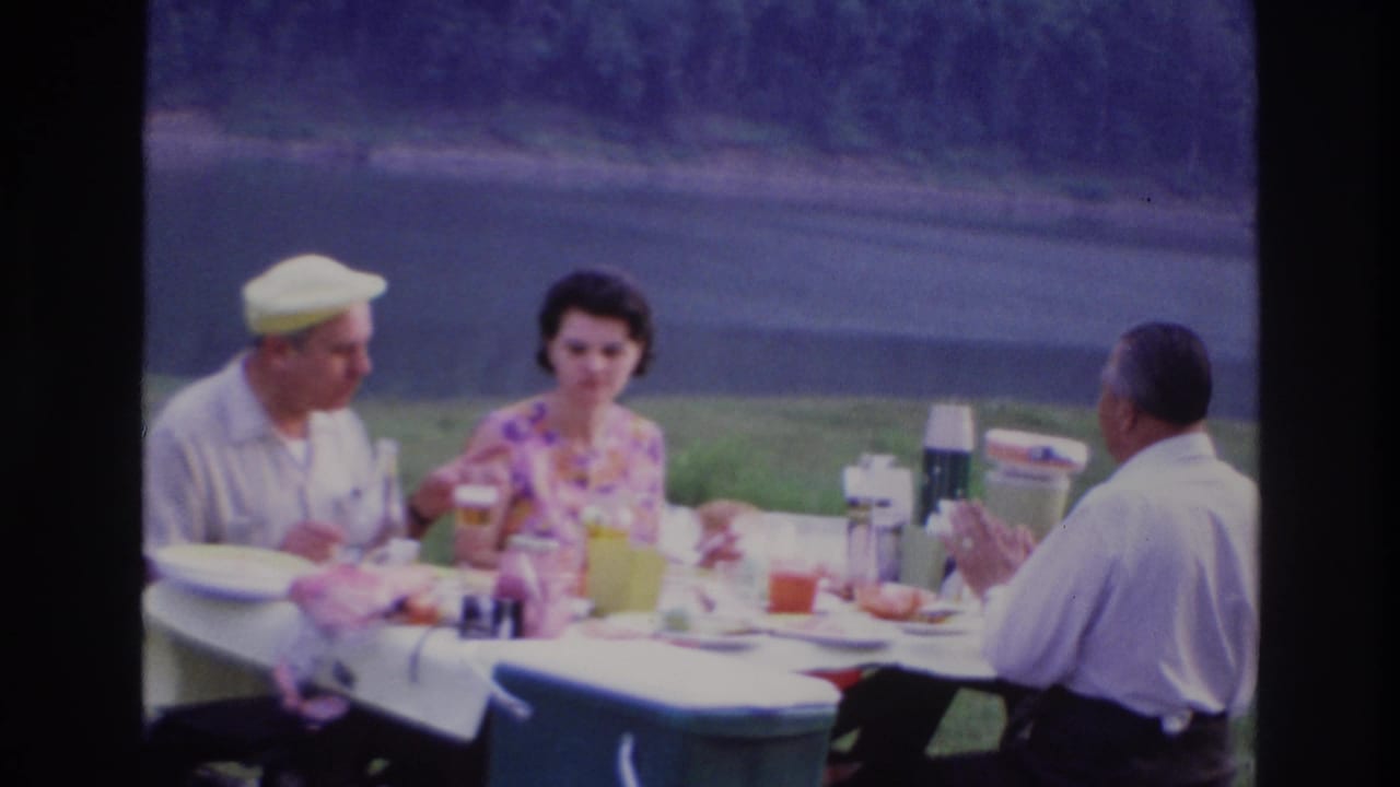 Three adults sit at an outdoor picnic table beside a body of water.