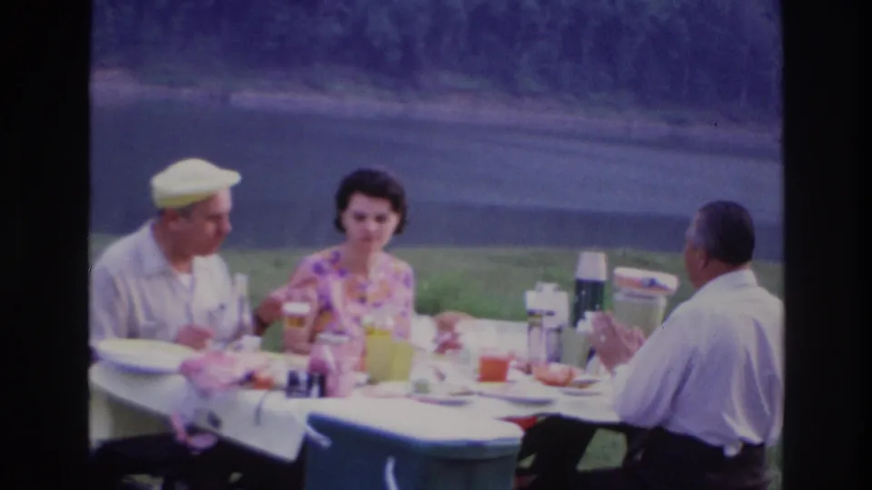 1967 Ottawa home-movie frame showing three adults at a picnic table with food, drinks, and thermoses near a lakeside lawn.