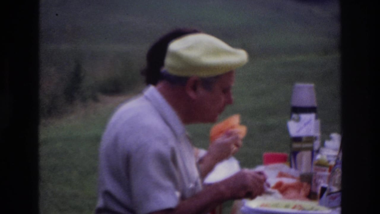 Close view of an adult man in a yellow cap eating at a picnic table, with thermoses and food visible in a grassy outdoor setting.