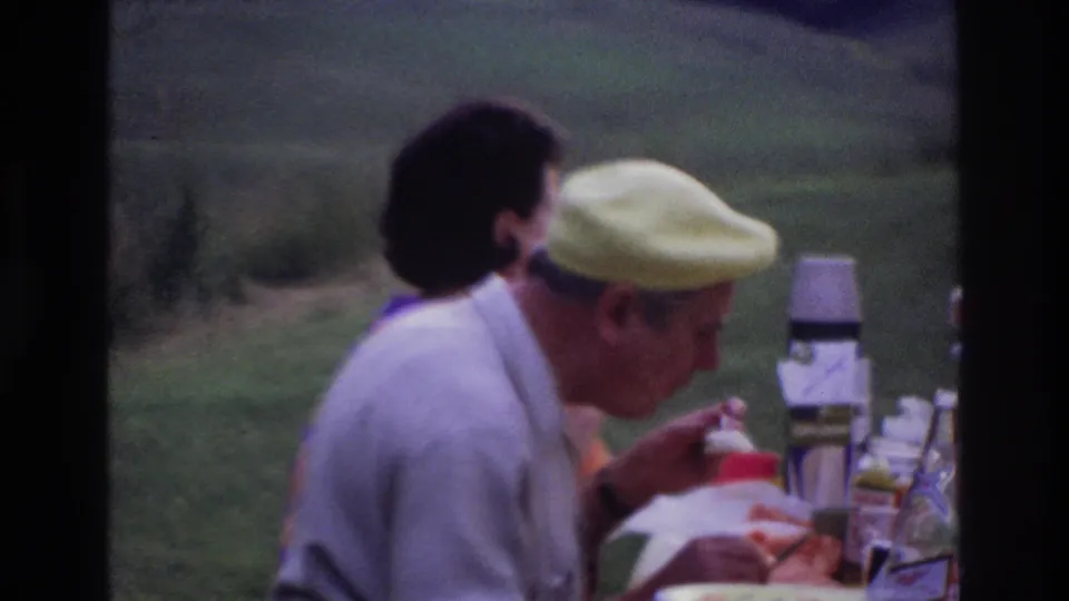 Home-movie frame of a man in a yellow cap leaning over food at a picnic table, with another adult partly visible behind him.