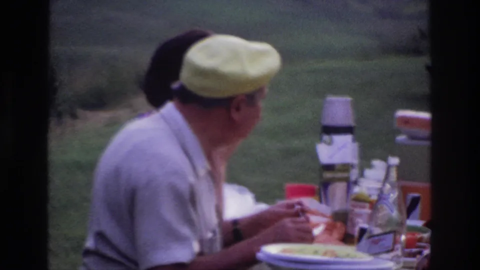 1967 Ottawa frame showing a man in a yellow cap turned toward another adult at a picnic table with plates, cups, and thermoses.