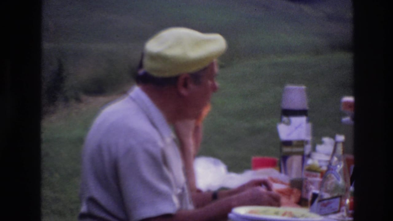 In 1967 Ottawa home-movie footage, a man in a yellow cap sits at an outdoor picnic table with food, cups, and a thermos nearby.