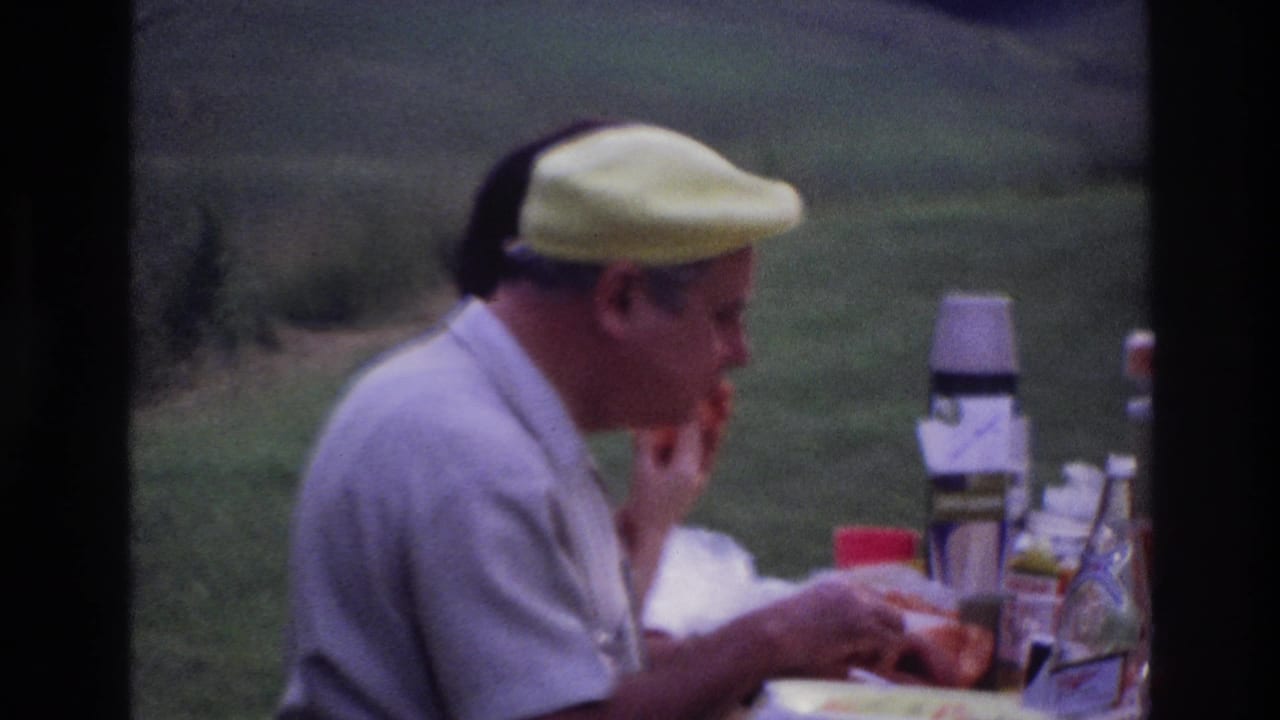 The seated man in a yellow cap eats during an outdoor picnic in Ottawa, with a thermos, paper cups, and food containers on the table.
