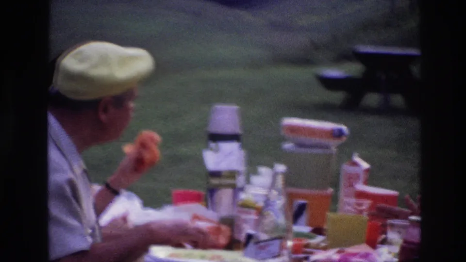 A 1967 Ottawa picnic scene shows the man holding food at the table, with cups, containers, grass, and a dark bench in the background.