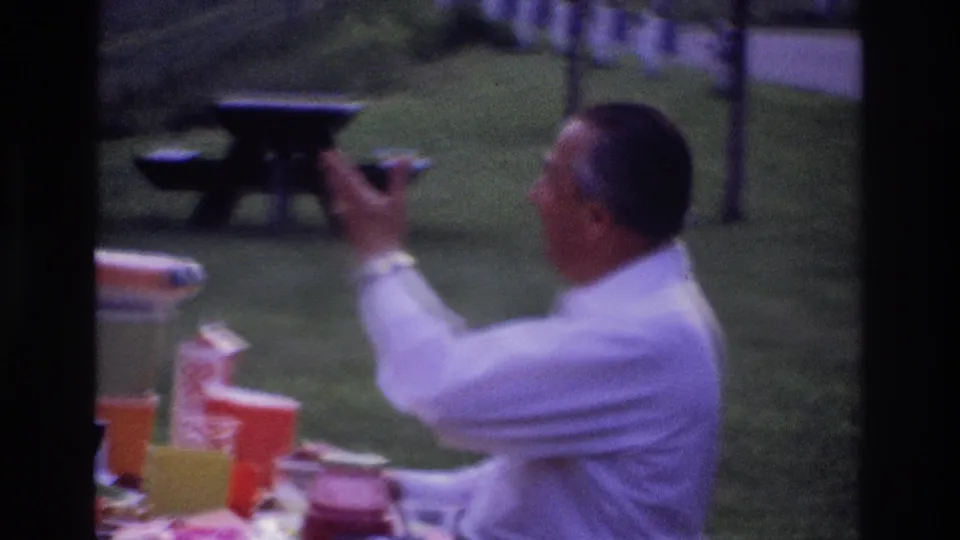 Another adult man in a white shirt gestures beside the picnic table during the Ottawa park meal, with cups, containers, grass, and a bench visible.