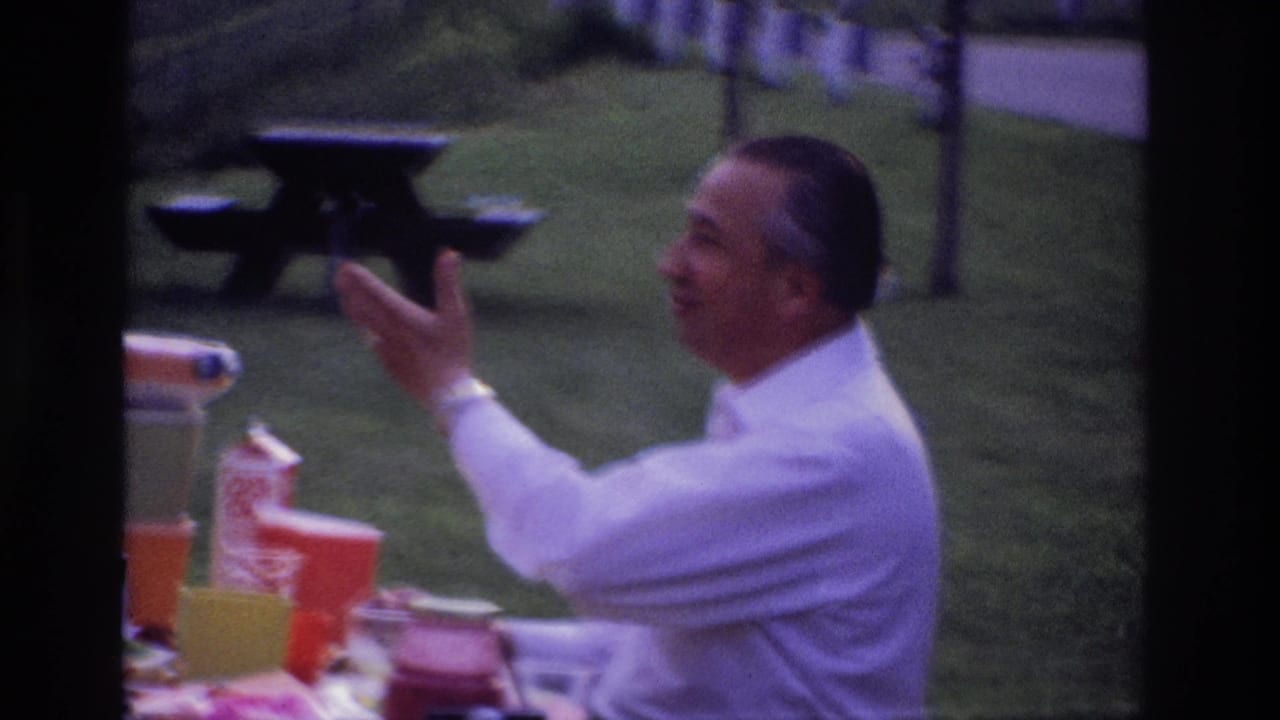 In 1967 Ottawa home-movie footage, a seated man in a white shirt gestures at a picnic table with cups and food containers nearby.