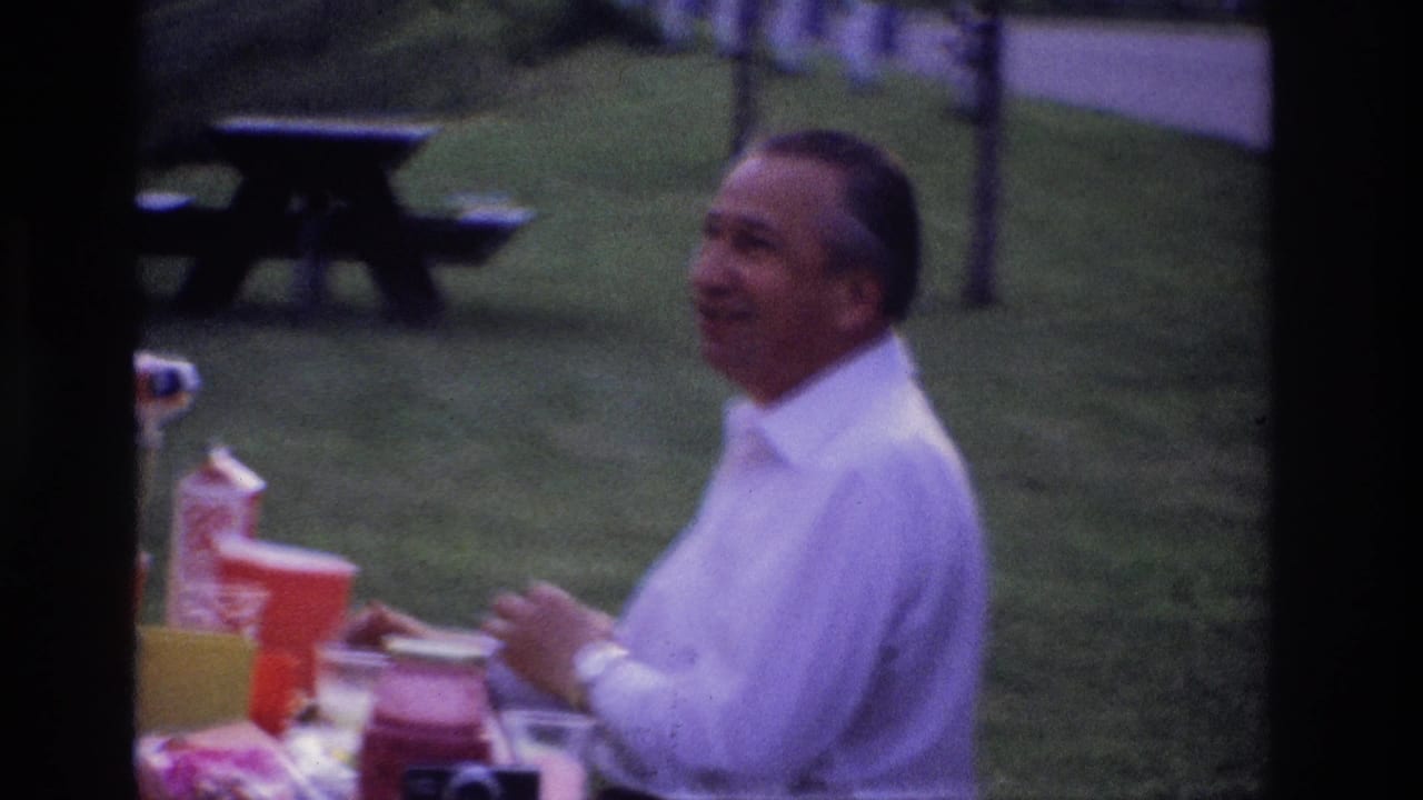 Man in a white shirt smiles at a picnic table with cups and food containers.