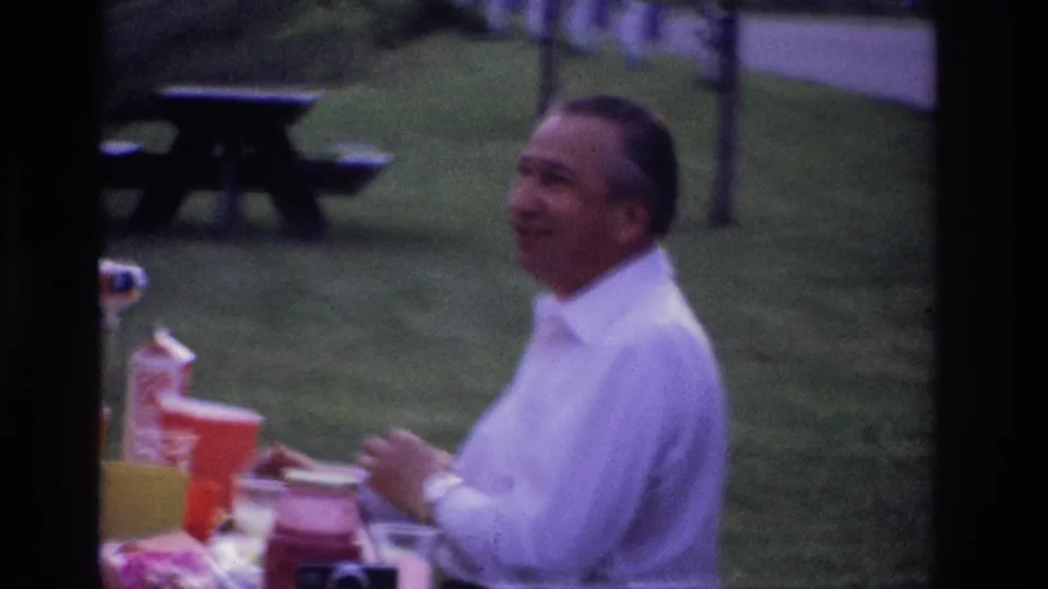 A smiling man in a white shirt sits at a picnic table in Ottawa, with colorful cups and containers visible in the park setting.