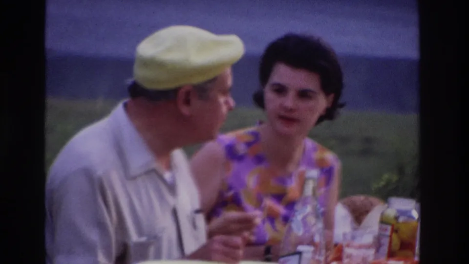 A man in a yellow cap and a woman in a purple-orange patterned dress converse at an Ottawa picnic table in 1967 home-movie footage.