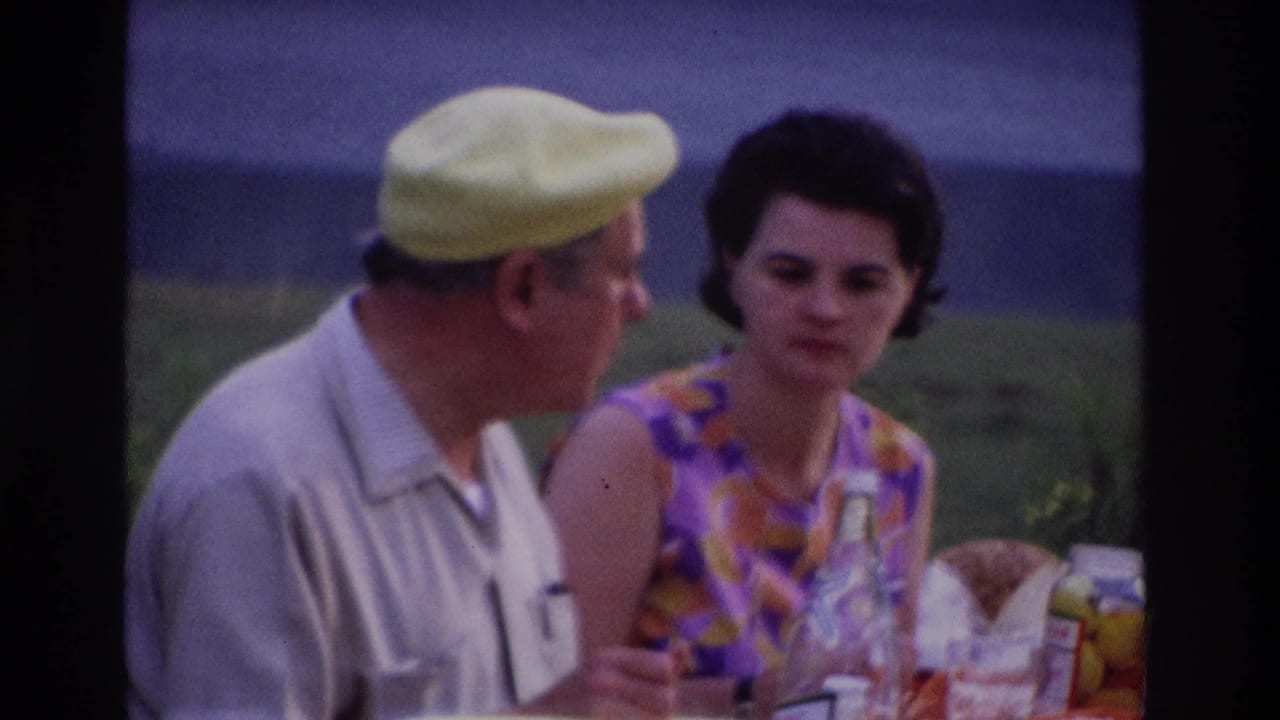 1967 Ottawa home-movie frame showing a man in a yellow beret and a woman in a purple-orange dress at a picnic table.