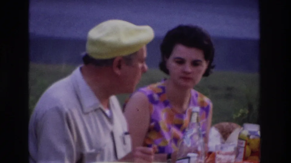 1967 Ottawa home-movie frame showing a man in a yellow beret and a woman in a purple-orange dress at a picnic table.
