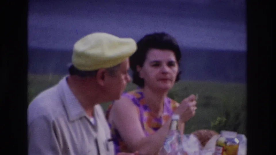 1967 Ottawa home-movie frame of a woman eating beside a man at a picnic table with food and drink items visible.