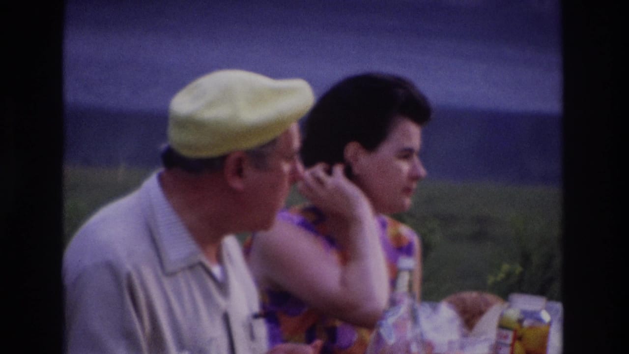 1967 Ottawa home-movie frame of a man in a yellow cap and a woman in a purple-orange dress seated at a lakeside picnic table.