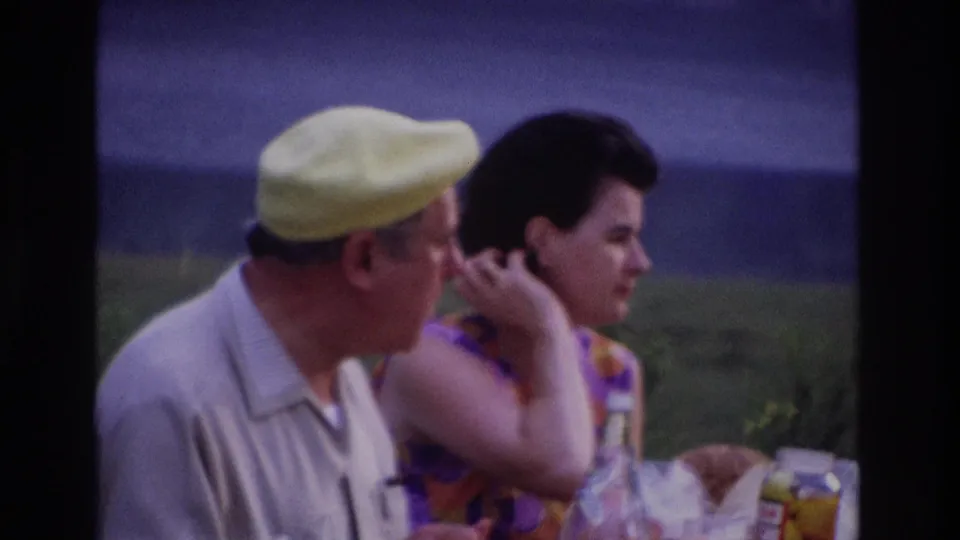 1967 Ottawa home-movie frame of a man in a yellow cap and a woman in a purple-orange dress seated at a lakeside picnic table.