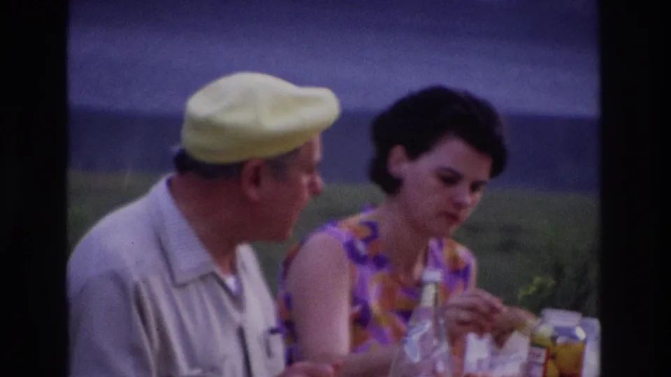1967 Ottawa lakeside picnic frame with a woman in a patterned dress and man in a yellow cap seated beside food and drink containers.