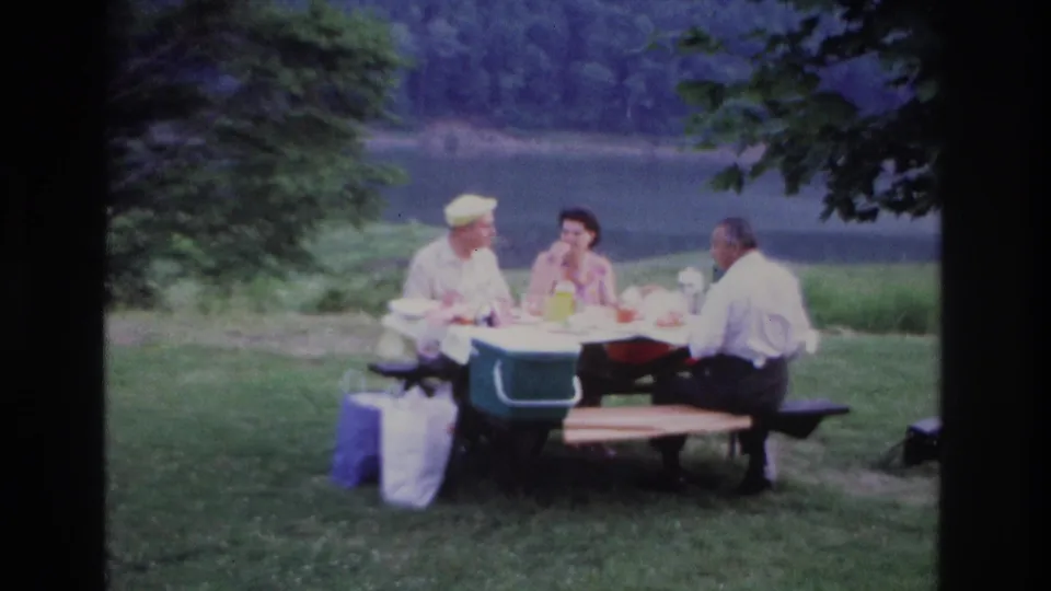 Wide 1967 Ottawa home-movie frame of three adults eating at a lakeside picnic table with a green cooler and trees nearby.
