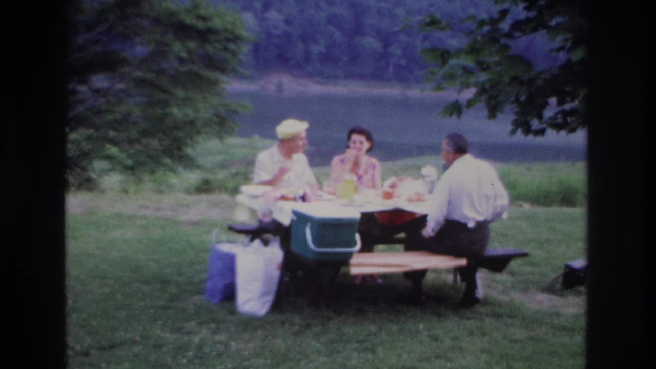 1967 Ottawa home-movie frame of three adults at a lakeside picnic table with a green cooler, bags, grass, trees, and a forested shoreline.