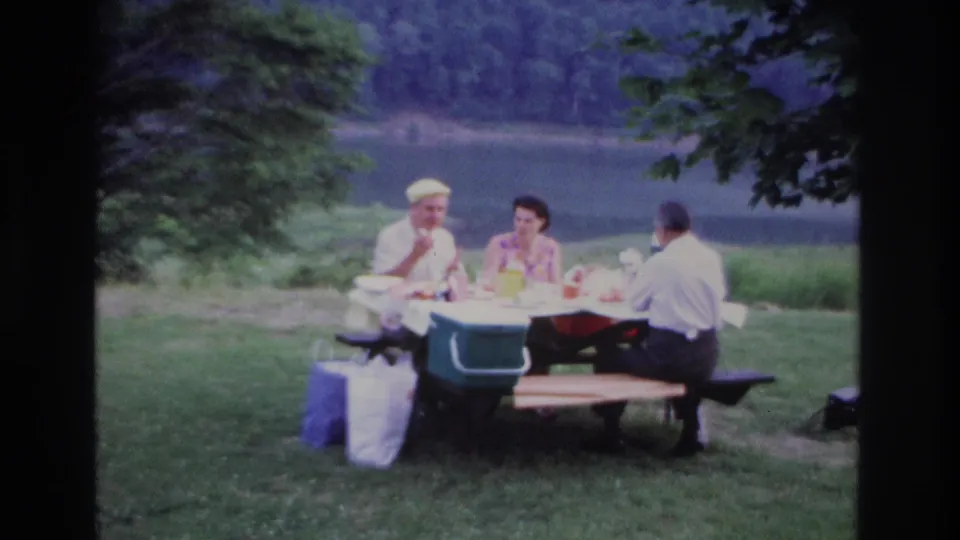 1967 Ottawa lakeside picnic frame showing three adults seated around lunch, a cooler at the table, bags on grass, and a calm lake behind them.