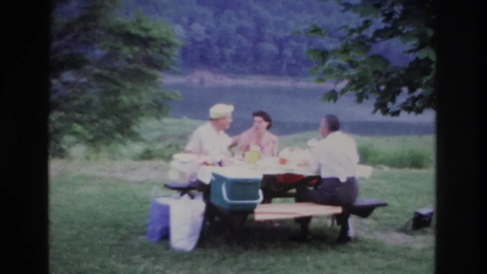 1967 Ottawa home-movie frame with a man in a light cap, a woman in a patterned dress, and another man at a picnic table near a lake.