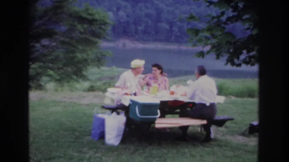 1967 Ottawa lakeside picnic frame showing three adults in conversation at a wooden table, with bags nearby, trees above, and water behind.