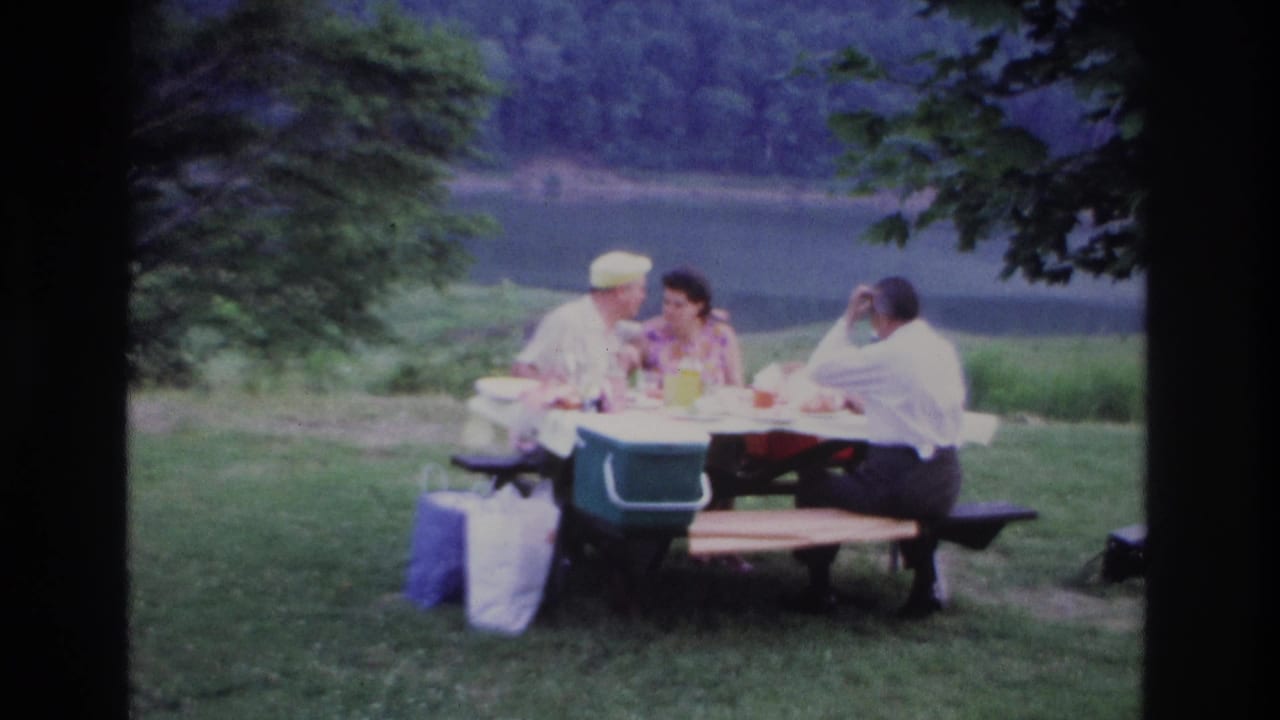Three adults sit at a picnic table beside a lake with bags on the grass.