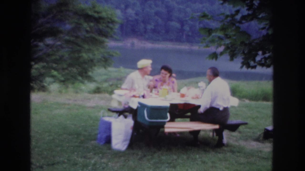 1967 Ottawa picnic scene with a man in a yellow cap, a woman, and another man seated at a wooden table near the water.