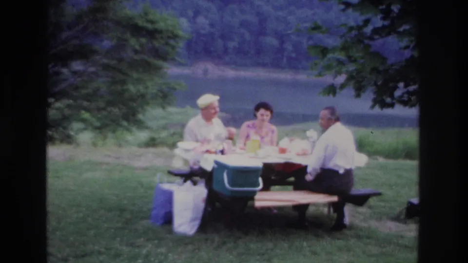 Three adults sit at a park picnic table in Ottawa in 1967, with picnic items on the table and calm water behind them.