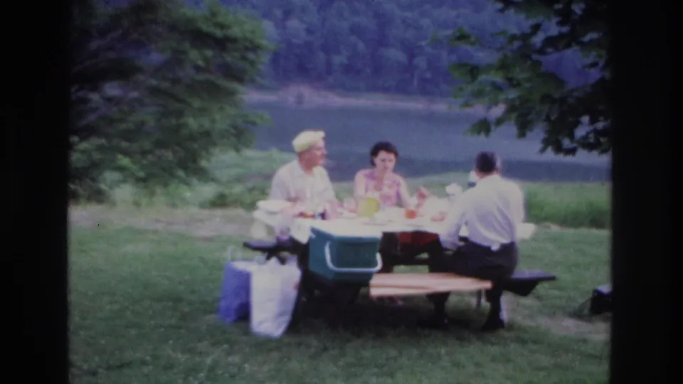 Final 1967 Ottawa frame of the lakeside picnic, with the seated group, green cooler, bags, grass, water, and wooded background.