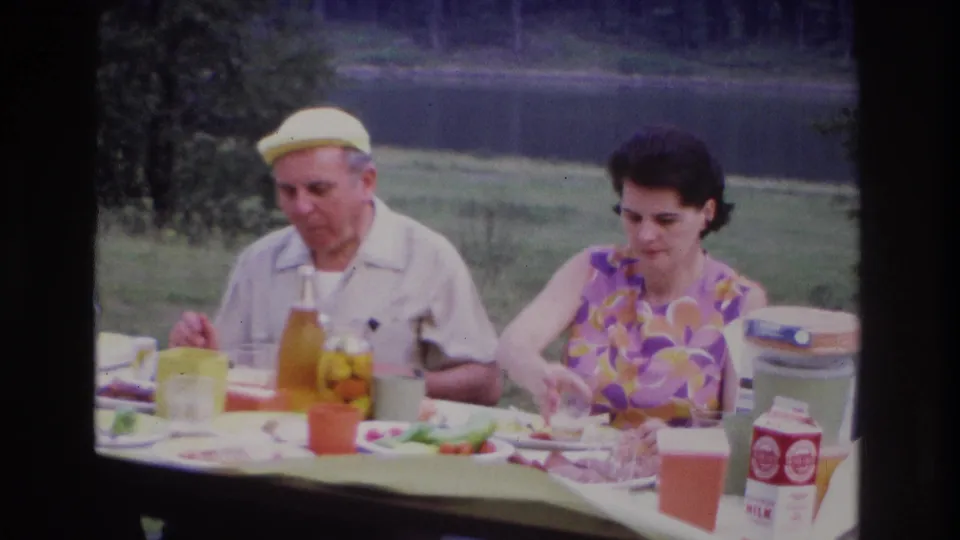 1967 Ottawa home-movie frame of two adults at a lakeside picnic table, with food, drink containers, and a cooler visible near the table.