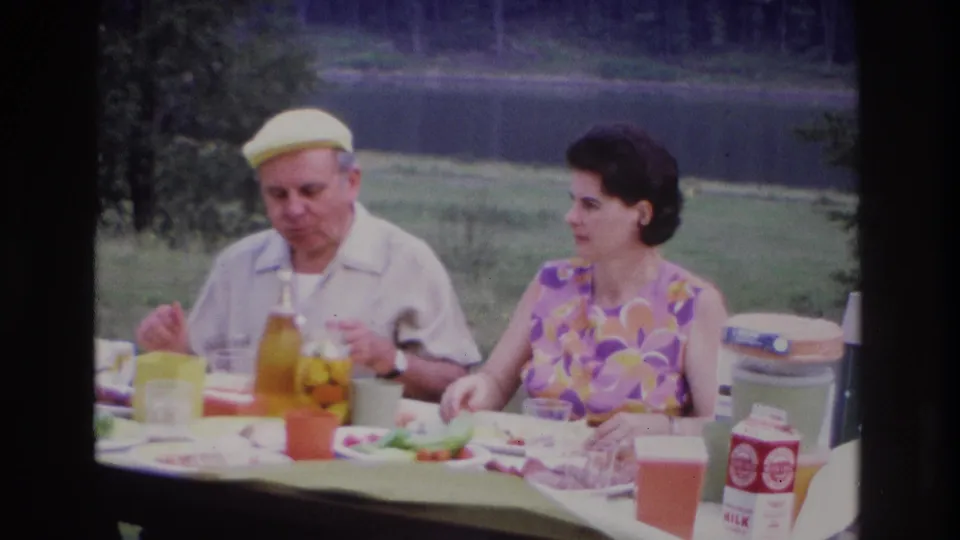 1967 Ottawa lakeside picnic frame with a man in a yellow cap and a woman in a floral dress seated among food, cups, cartons, and a cooler.