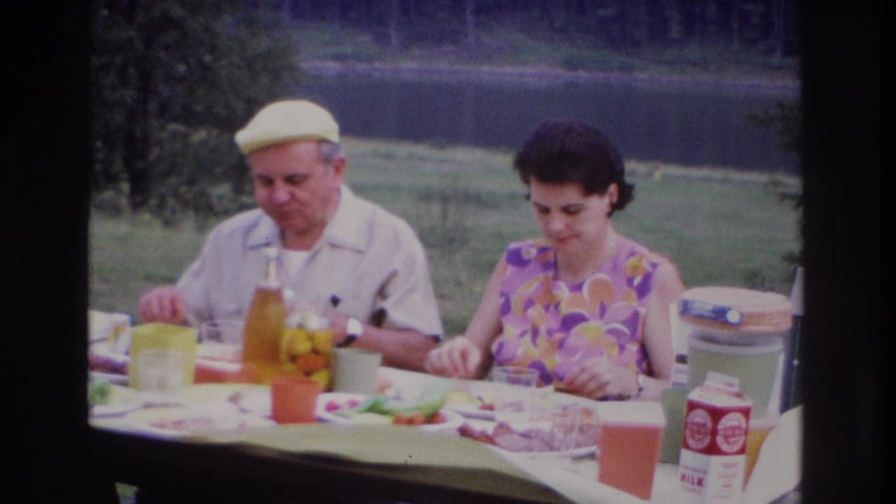 In Ottawa 1967 home-movie footage, a man in a yellow cap and a woman in a floral dress sit at an outdoor picnic table near a pond.