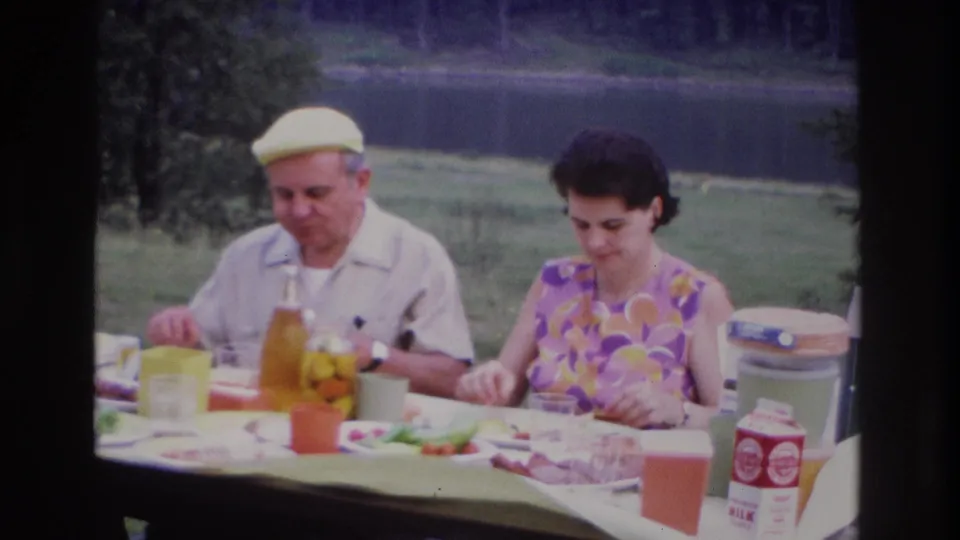In Ottawa 1967 home-movie footage, a man in a yellow cap and a woman in a floral dress sit at an outdoor picnic table near a pond.