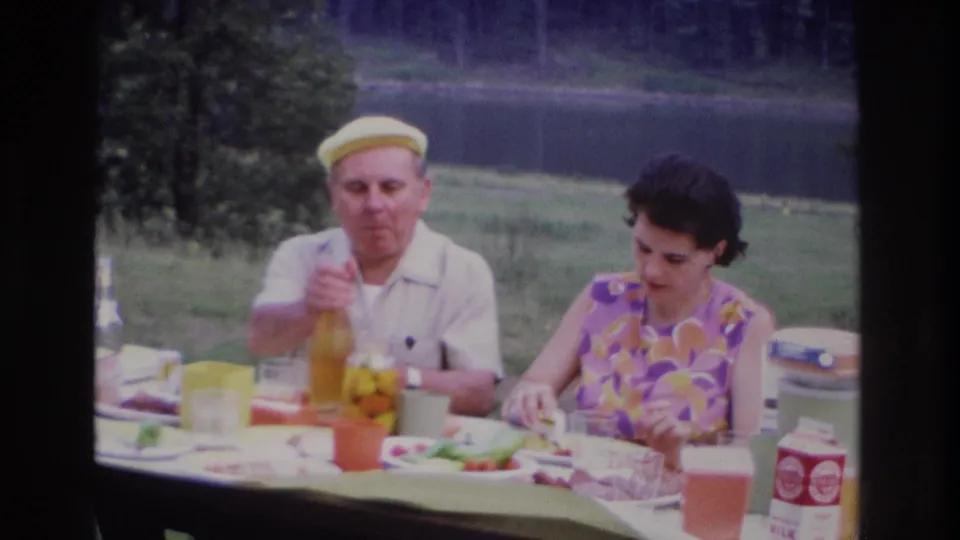 The man in a yellow cap reaches for the bottle as the woman looks down at food on the 1967 Ottawa picnic table near the pond.
