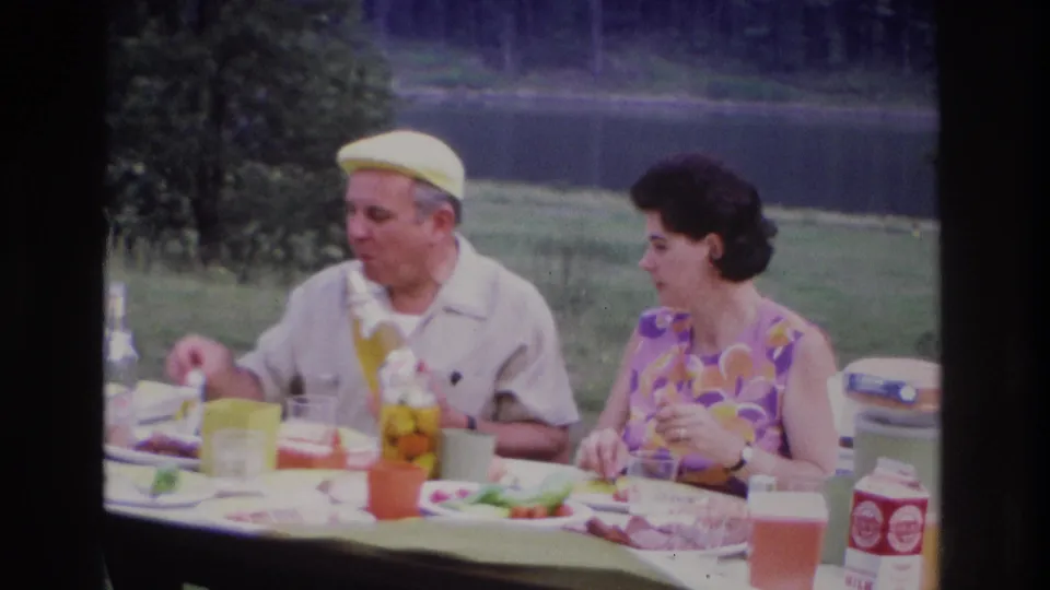 1967 Ottawa home-movie frame of a seated couple pausing over a picnic meal, with lake and trees behind them.
