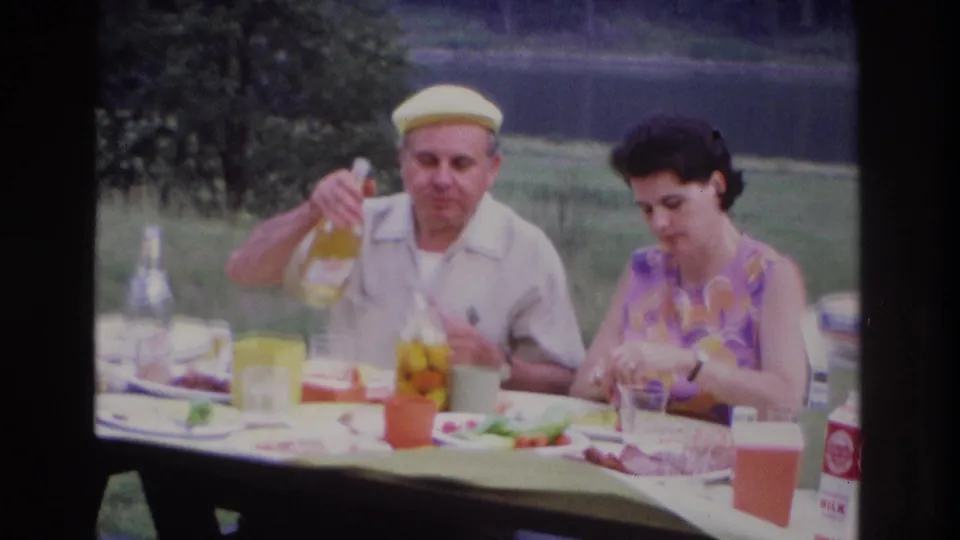 1967 Ottawa lakeside picnic frame showing the man holding a bottle and the woman eating at a table set with food and drink items.