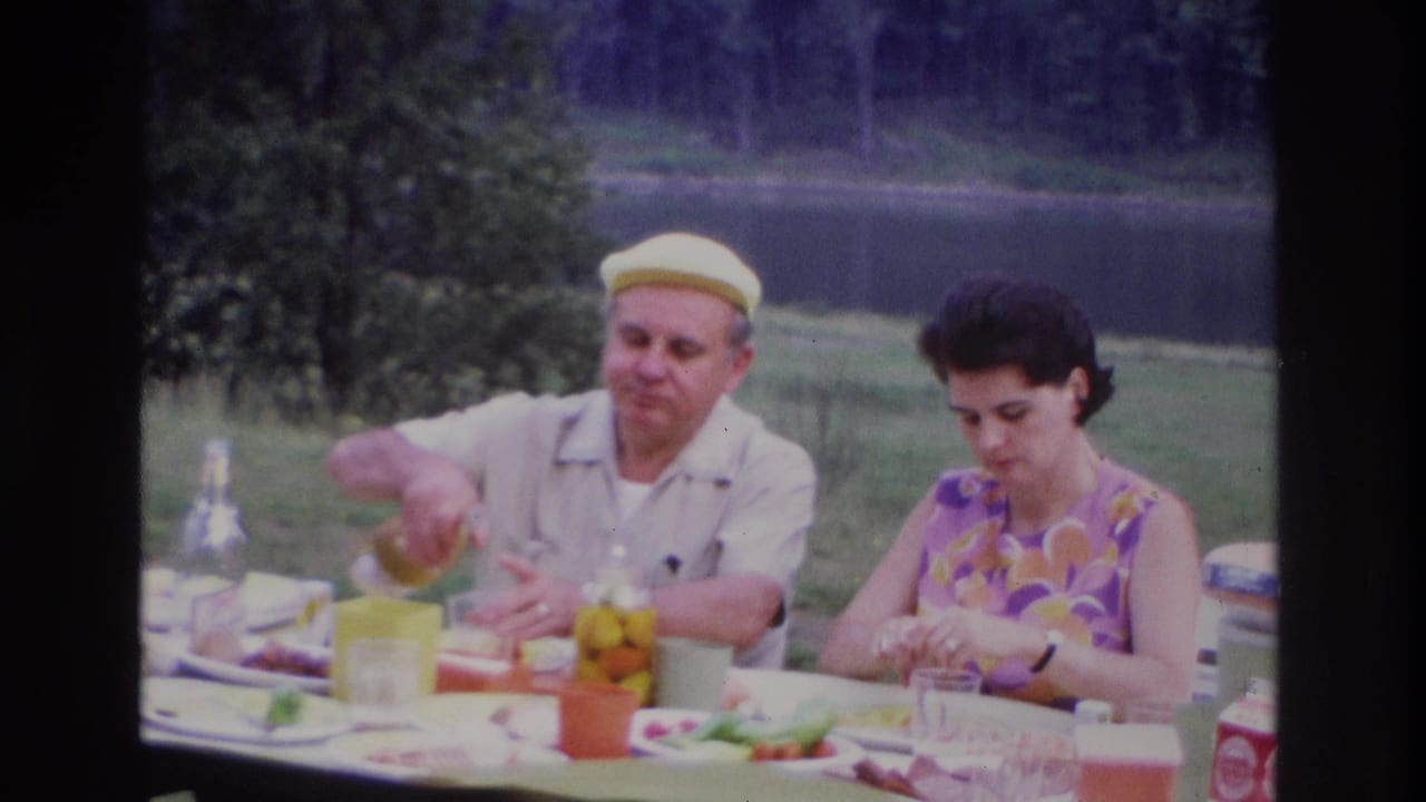 1967 Ottawa home-movie frame of a man pouring a drink while a woman sits at a lakeside picnic table with food and cups.