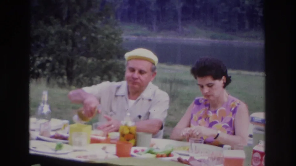 1967 Ottawa home-movie frame of a man pouring a drink while a woman sits at a lakeside picnic table with food and cups.