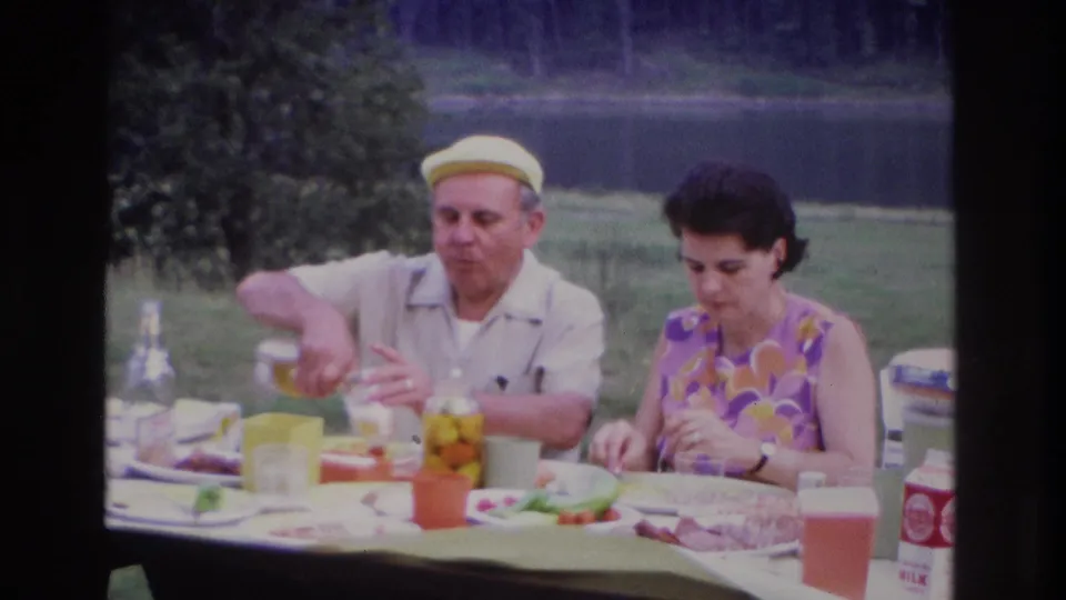 1967 Ottawa home-movie frame of two adults at a picnic table with food and drinks on a grassy lakeside setting.