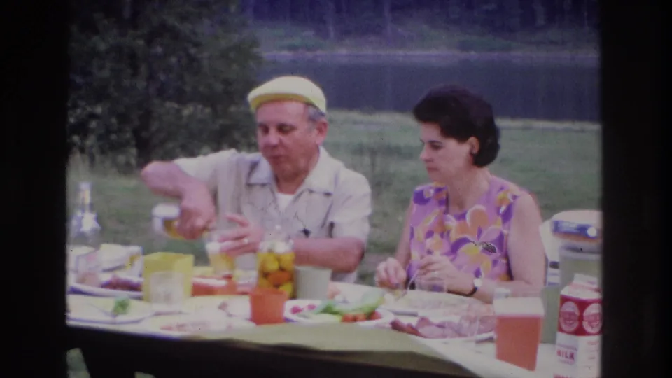1967 Ottawa lakeside picnic frame with a man pouring a drink and a woman looking toward him at a table with food.