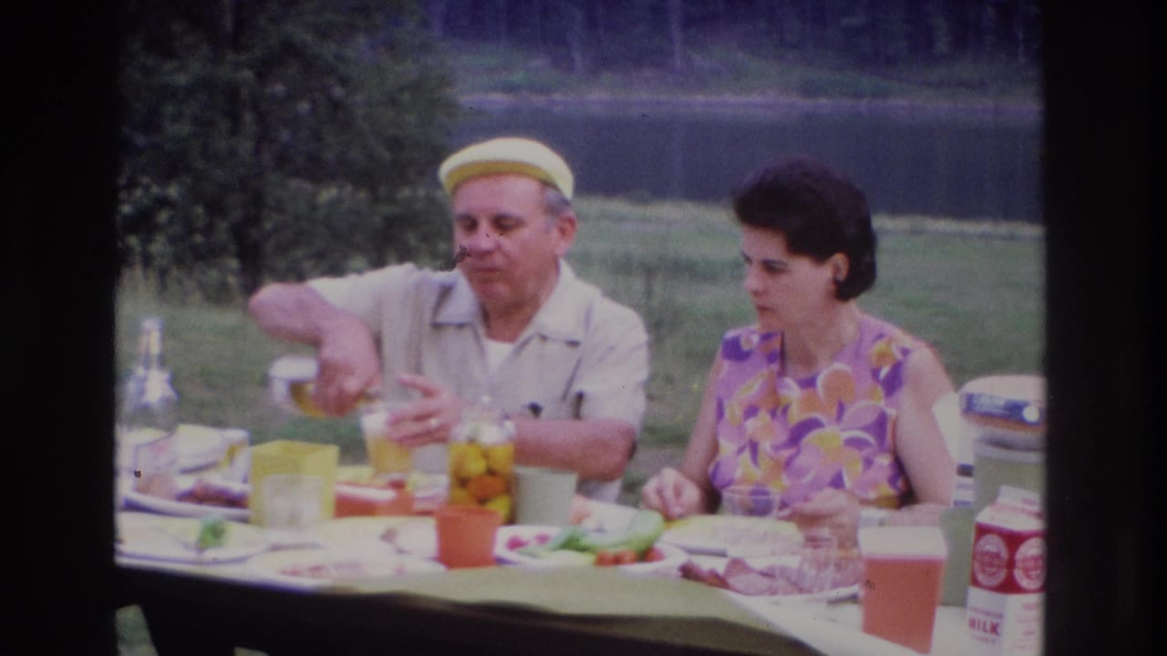 Man in a yellow cap pours a drink at a picnic table while a woman sits beside him.
