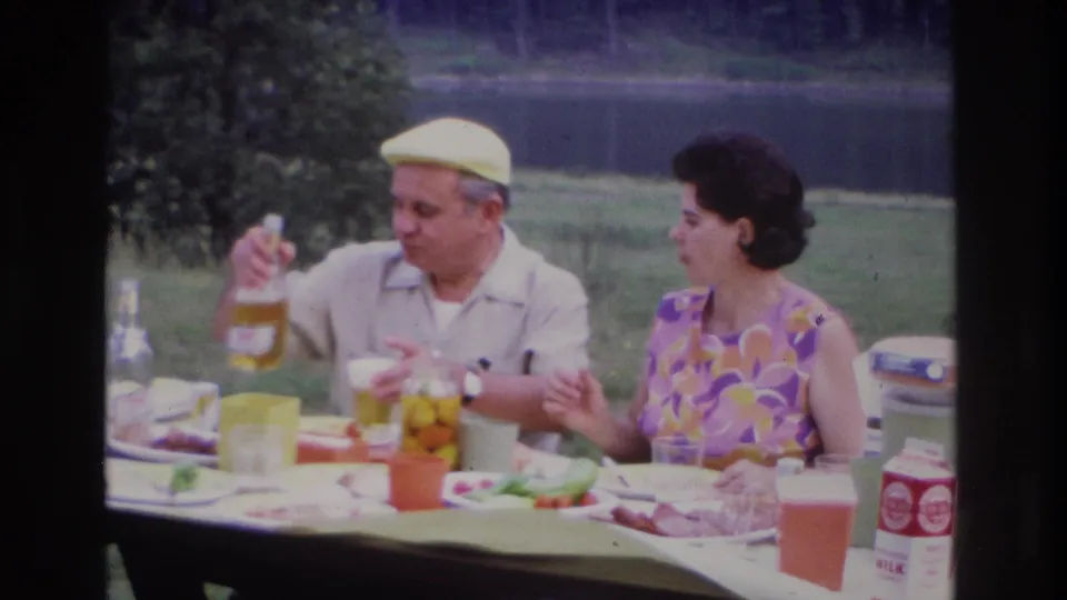 1967 Ottawa lakeside picnic frame with a man holding a bottle and a woman gesturing near plates, glasses, and cartons.