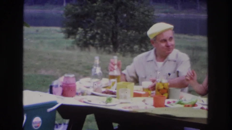 1967 Ottawa home-movie frame of a man at a lakeside picnic table with a bottle, food, glasses, and cartons visible.