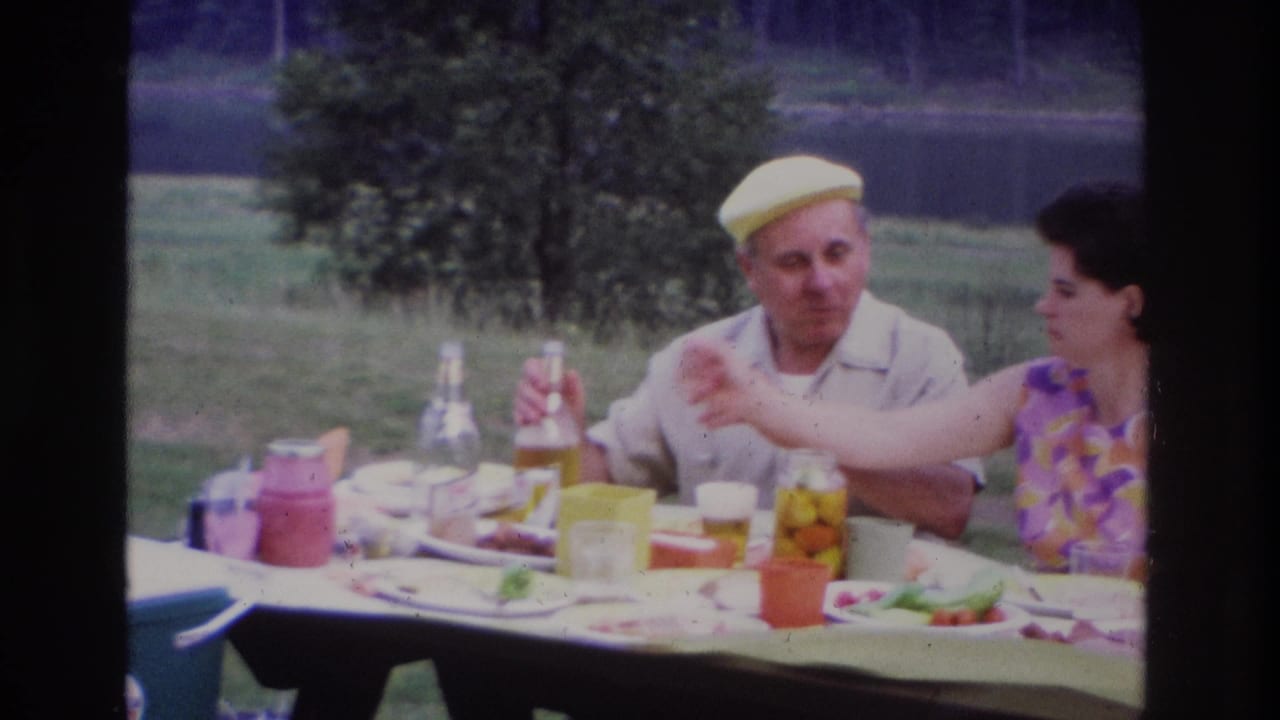 1967 Ottawa home-movie frame of an elderly couple at a lakeside picnic table with jars, cups, cartons, and food visible.