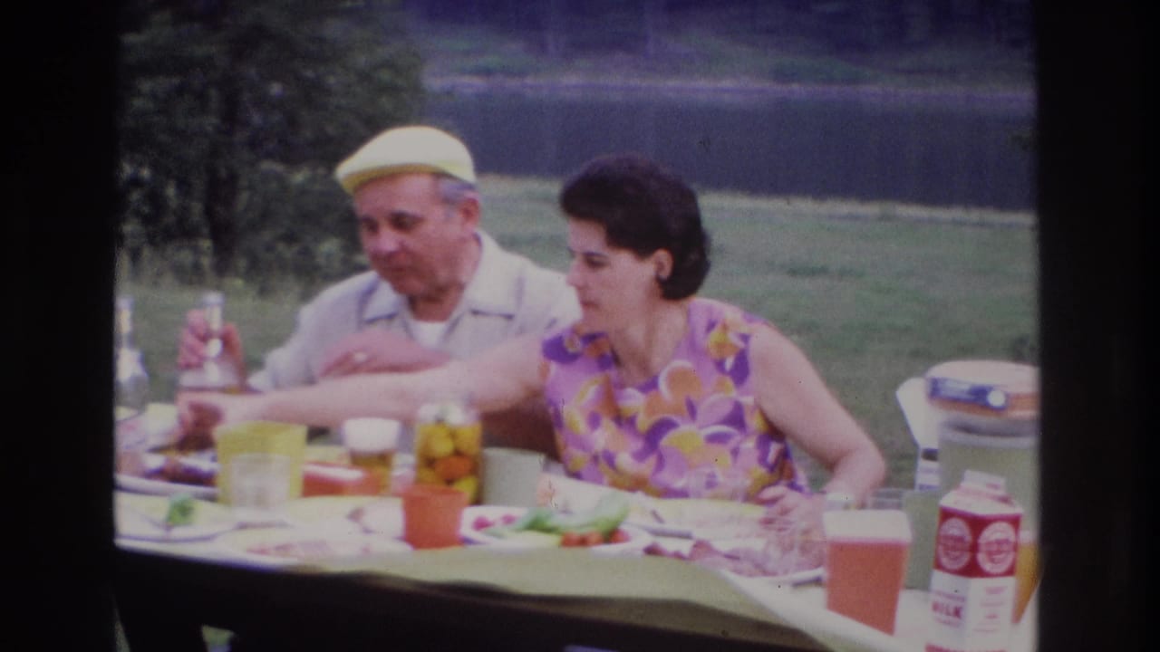1967 Ottawa lakeside picnic frame showing a woman reaching across the table as an elderly man watches, with food and cartons visible.